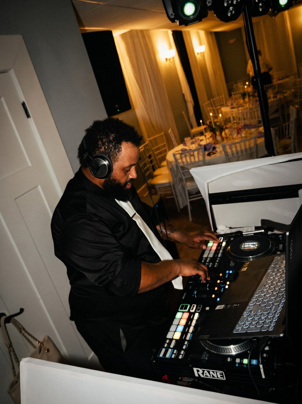 DJ in headphones mixing behind the booth as moving-head lights wash a reception room.