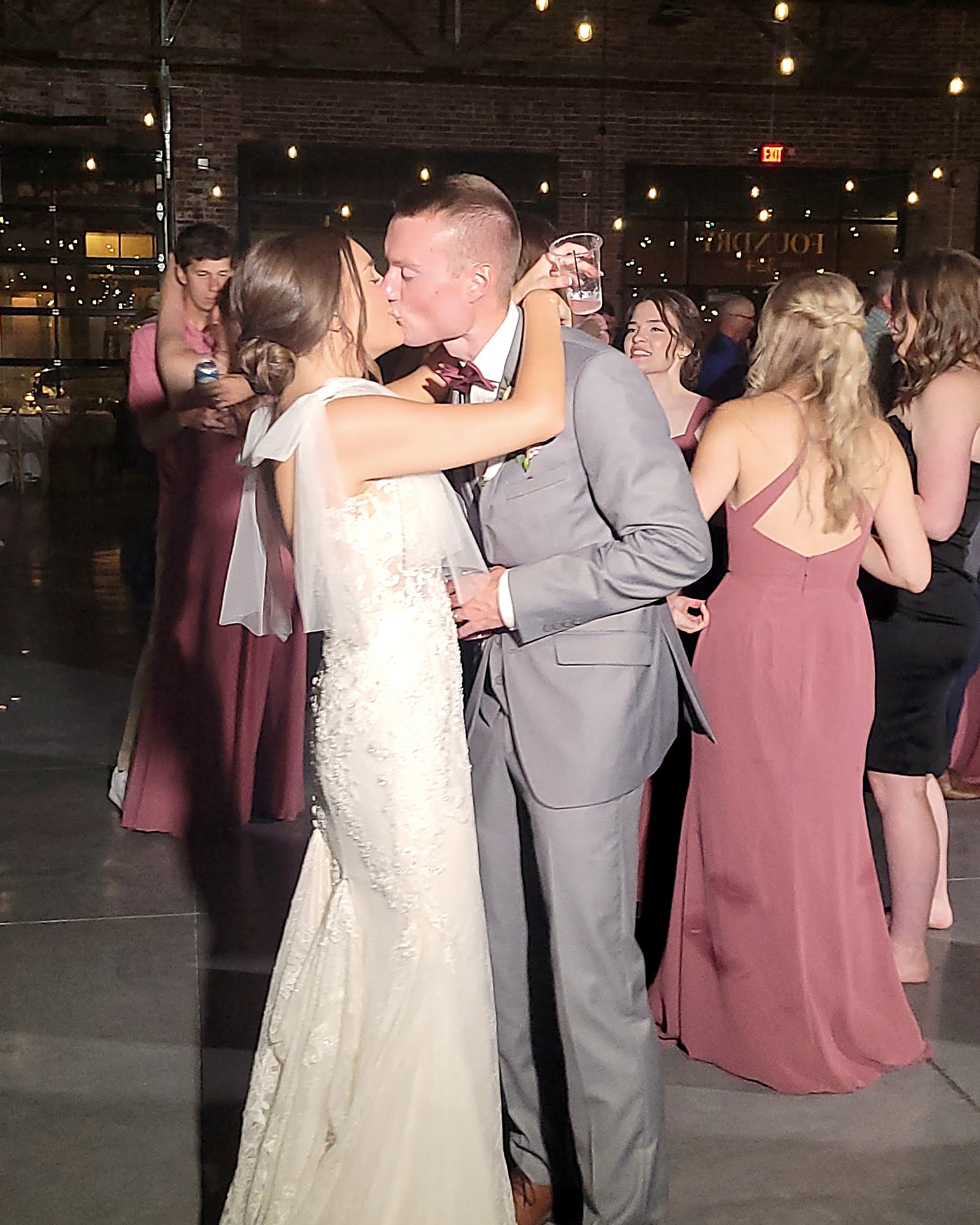 Bride and groom kissing during their first dance under warm string lights, wedding party around them.